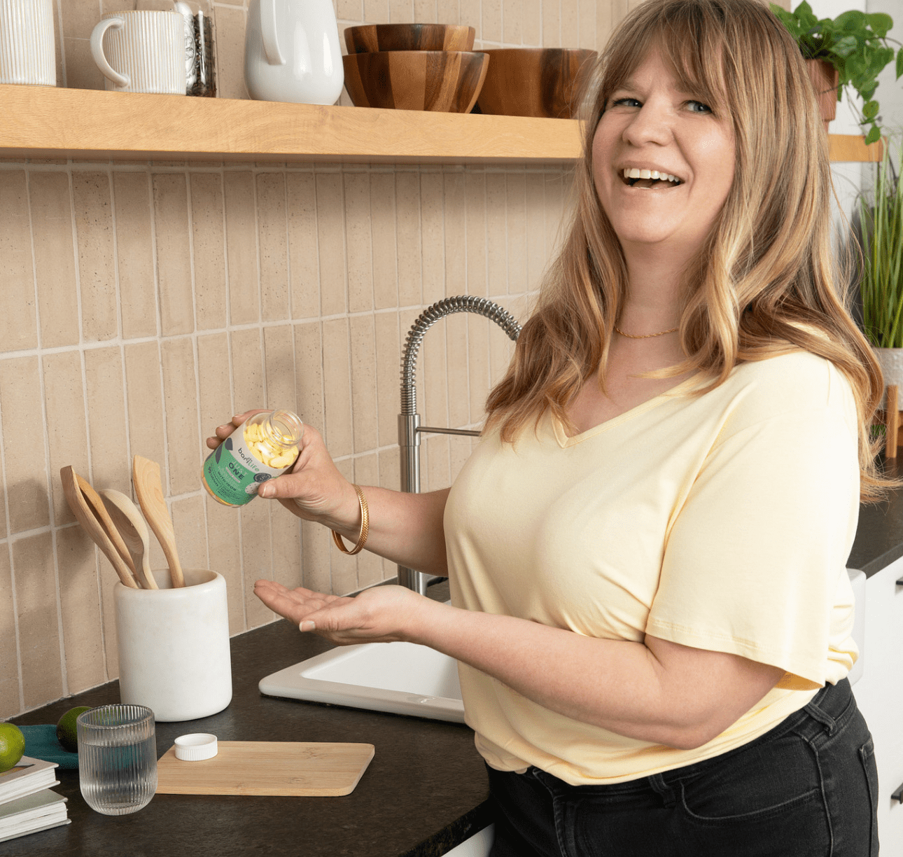 Smiling woman in a kitchen, holding a jar of kitchen ingredient and demonstrating its use on her hand.